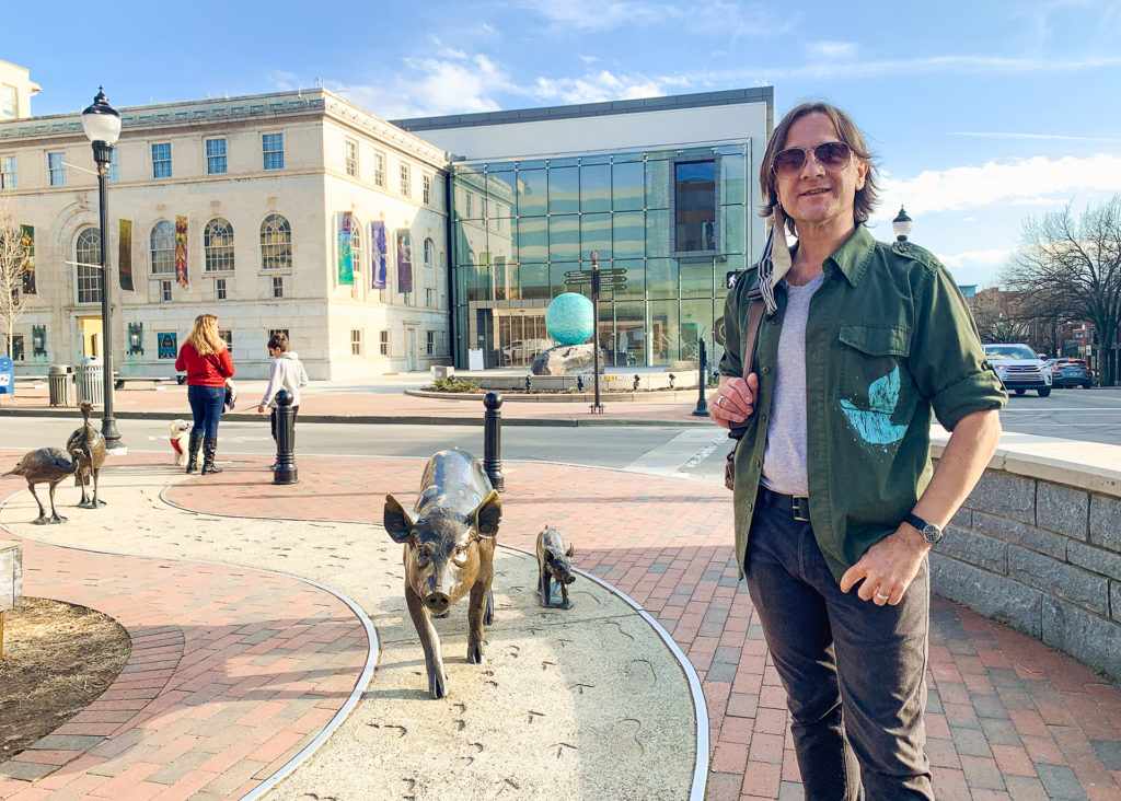 Stephen stands in downtown Asheville, NC near the art museum and in front of bronze statues of pigs and turkeys