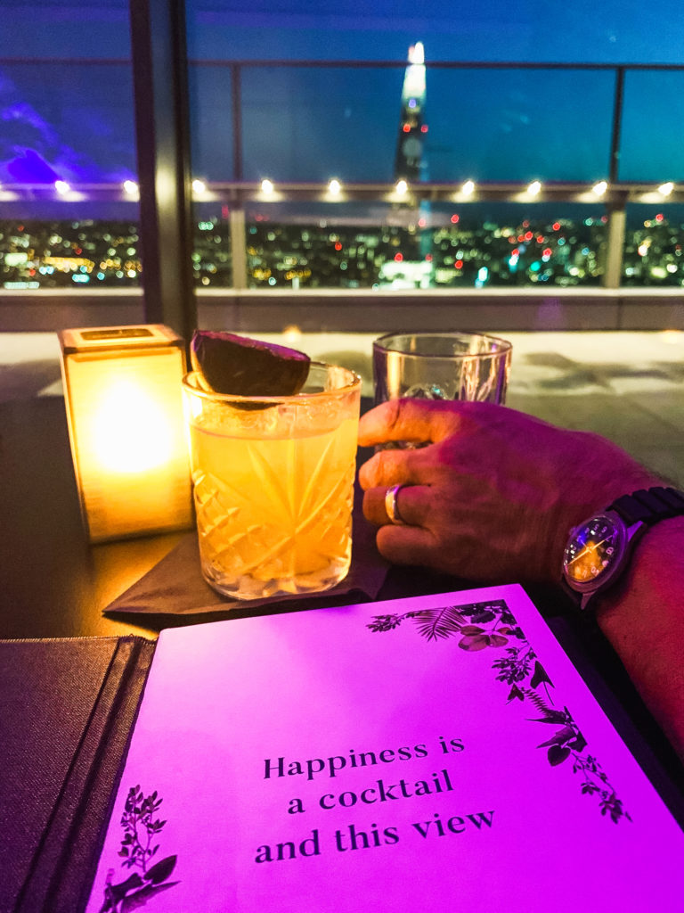 The menu, a candle, a man's hand holding a drink, and a nighttime view of London from Sky Garden