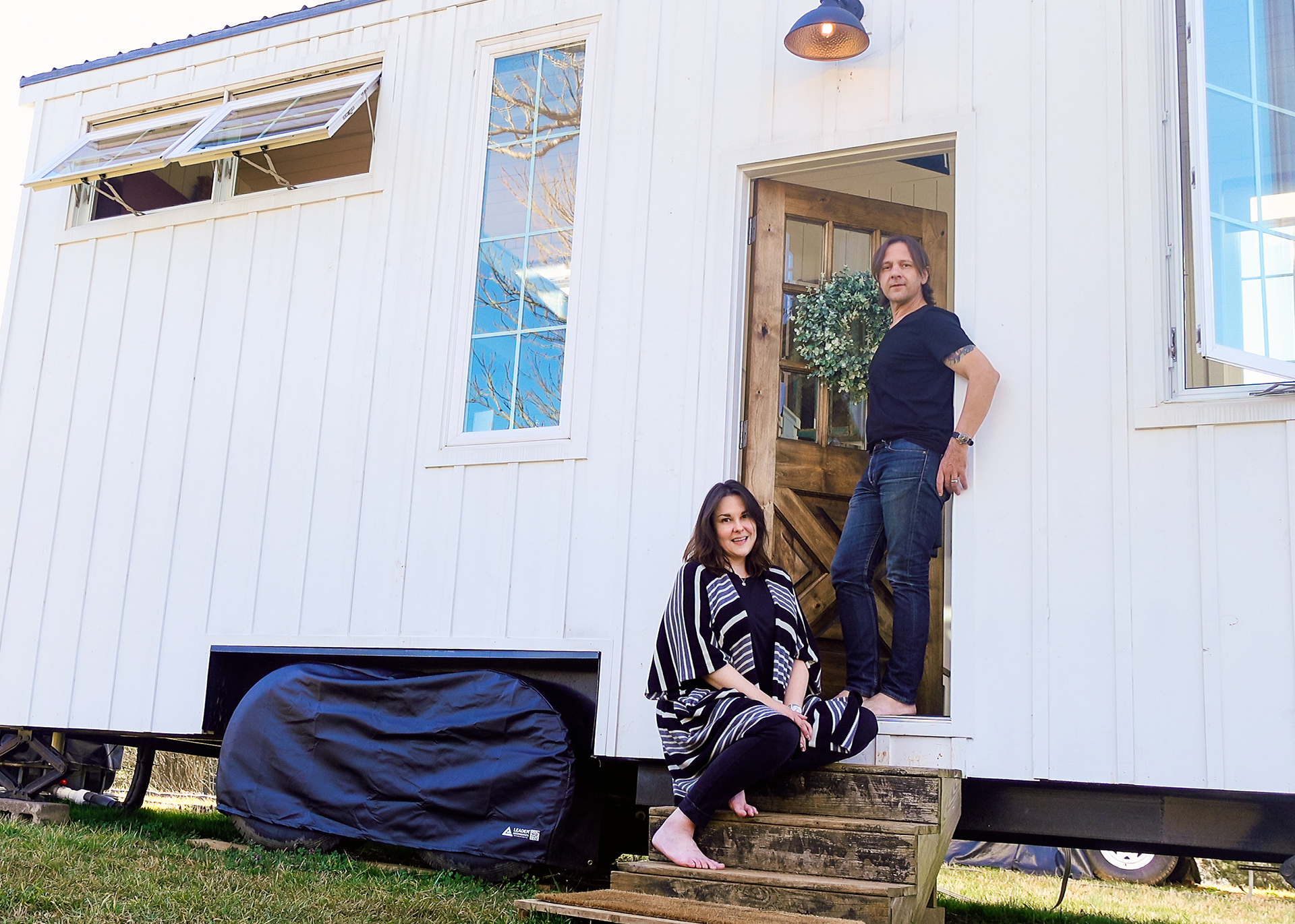 Andie and Stephen pose at the front entrance to a white farm-style tiny house on a farm