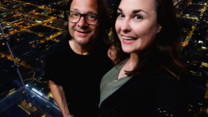 a man and woman stand in the glass cube on the 103rd floor of Chicago's Willis Tower, with a night view of the city lights below them. it's their 1-year vlogging anniversary
