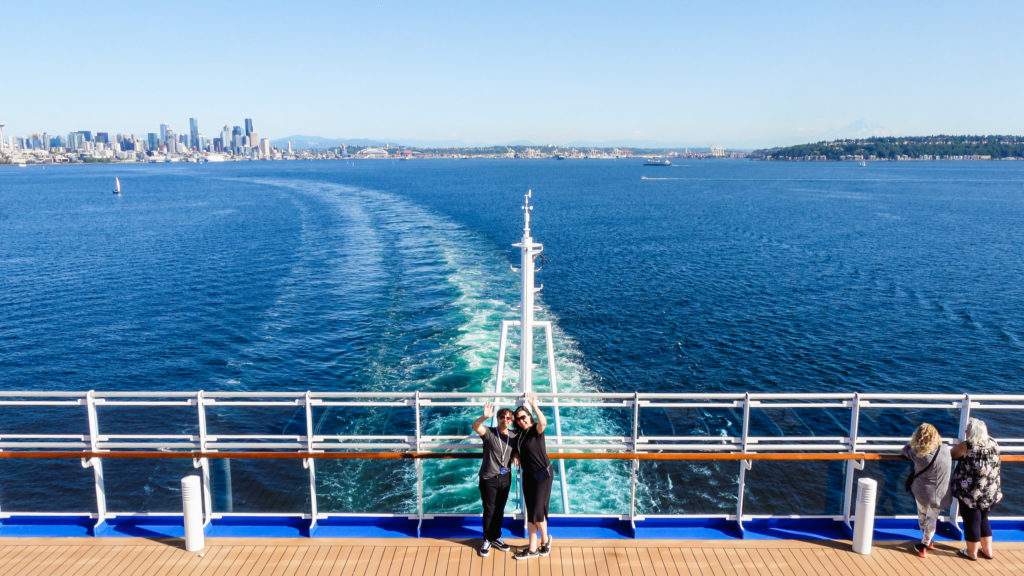 Stephen and Andie wave to the camera while standing on deck of a cruise ship with the ship's wake and the city of Seattle behind them