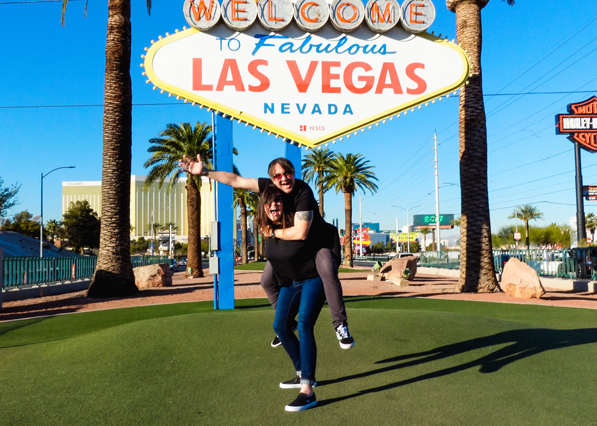 man and woman posing in front of the iconic "Welcome to Fabulous Las Vegas" sign in Las Vegas. The woman appears to be giving the man a piggyback ride.