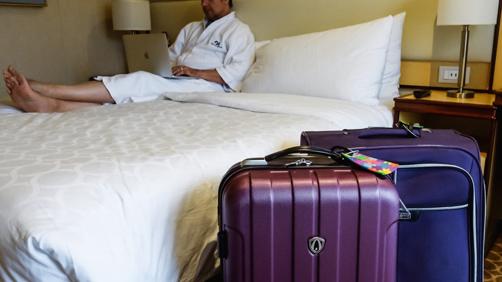 man sitting in bathrobe on bed in his cruise ship cabin with suitcases next to the bed