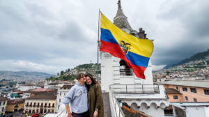 man and woman on top of a church overlooking Quito, Ecuador. The Ecuador flag waves behind them