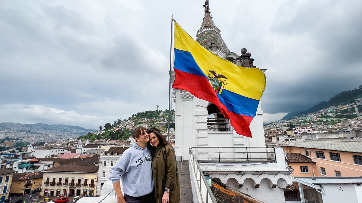 man and woman on top of a church overlooking Quito, Ecuador. The Ecuador flag waves behind them
