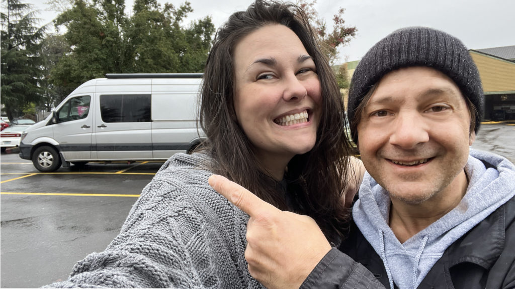 man and woman smiling, pointing behind them to their new-to-them silver Sprinter campervan. mission to see all 50 states.
