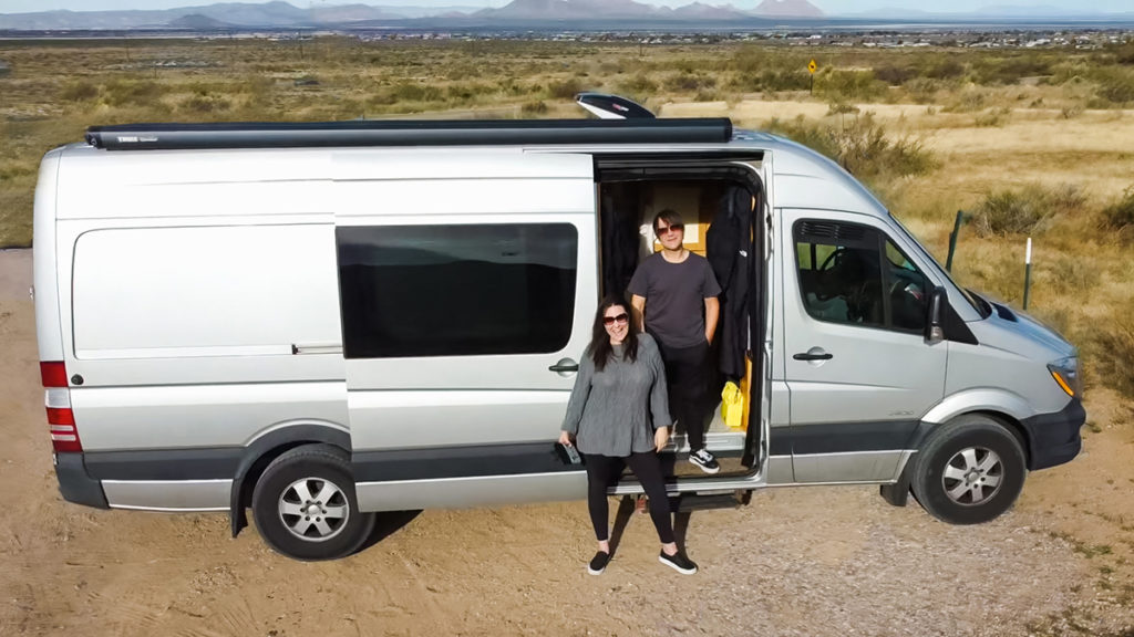 aerial drone view of man standing inside and a woman outside of the open sliding door of their silver Sprinter camper van. parked in an open land in Las Cruces, New Mexico. they give 5 van life lessons in this post.