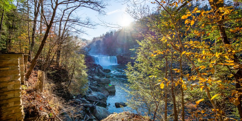 a large, distant waterfall in Cumberland Falls, Kentucky with blue water, green trees, and the sun peeking just over the hill behind it.