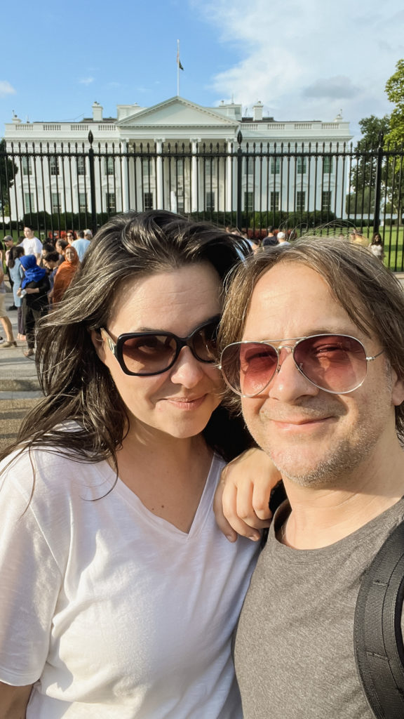 a smiling woman and man pose in front of the White House in Washington, DC