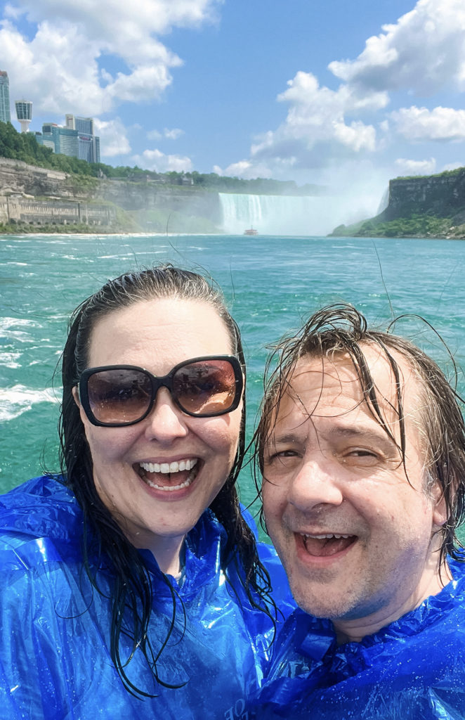 a smiling woman and man in blue plastic ponchos, with soaking wet hair and faces and a large waterfall at Niagara Falls behind them