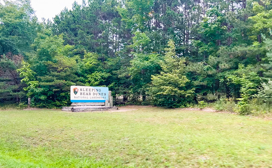 photo of the Sleeping Bear Dunes National Lakeshore sign on green grass surrounded by many various lush green trees, near one of the entrances to the Northern Michigan park.