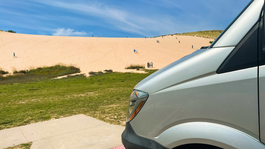 view of the Dune Climb at Sleeping Bear Dunes National Lakeshore in Northern Michigan, with the nose of a silver Sprinter camper van in the foreground. various people are scattered, climbing the dune.