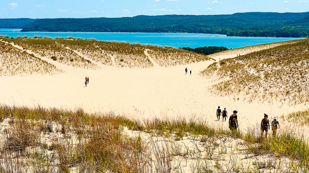 view of multiple people hiking across the sand dunes and trails of Sleeping Bear Dunes National Lakeshore in Northern Michigan. Overlooking the aqua blue waters of Glen Lake in the distance on a blue sky afternoon.