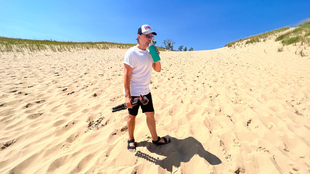 man standing in the sand drinking water from a reusable water bottle, holding a camera, while doing the Dune Climb at Sleeping Bear Dunes National Lakeshore in Northern Michigan on a blue sky afternoon.