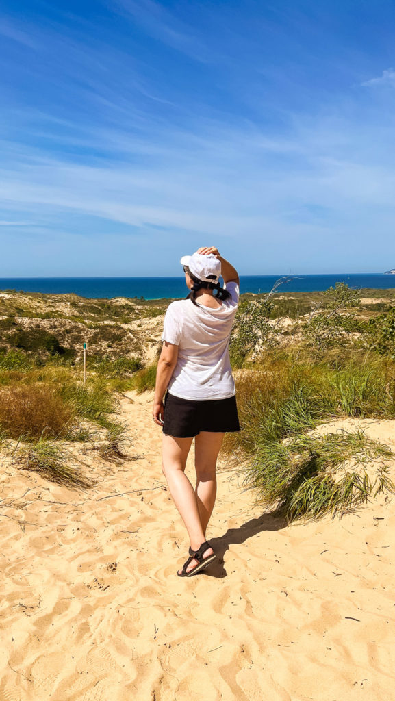 man standing in the sand drinking water from a reusable water bottle, holding a camera, while doing the Dune Climb at Sleeping Bear Dunes National Lakeshore in Northern Michigan on a blue sky afternoon.