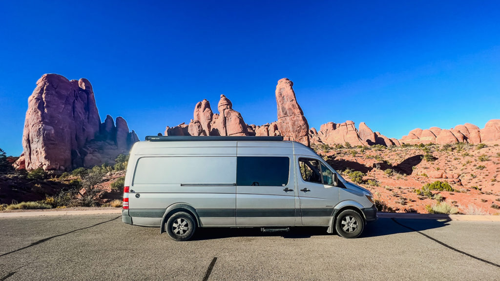 a silver Sprinter camper van is parked in front of one of the many impressive rock formations in Arches National Park in Utah. A perfectly blue sky is in the background.