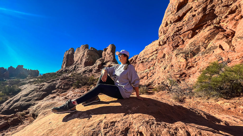 a woman sits atop an impressive rock formation, smiling and gazing out at the landscape of Arches National Park in Utah.