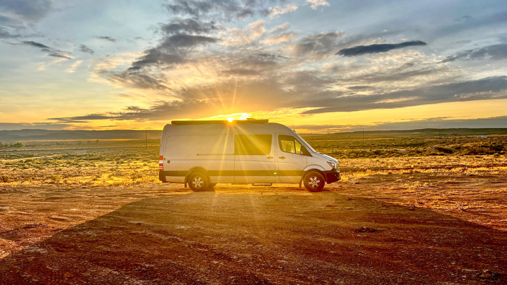 a silver Sprinter camper van is parked in an vast, open boondocking location in Moab, Utah with the sun setting behind the van. the sunset appears to be blasting out from the top of the van.