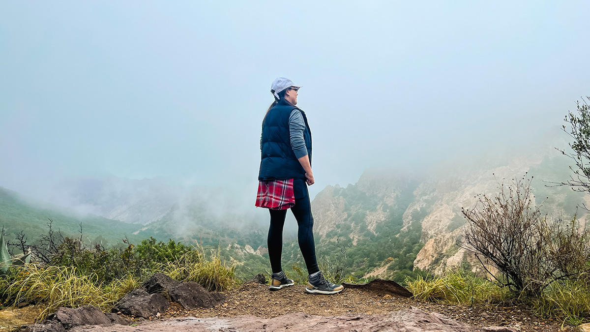 A woman standing in a very foggy, cloudy landscape in Big Bend National Park. She is taking a break from hiking and overlooking a canyon which is hardly visible in the fog. Some grass, dirt, and rocks are around her feet, and she is dressed for an autumn day, wearing a cap, long sleeved shirt, puffer vest, leggings, and hiking shoes