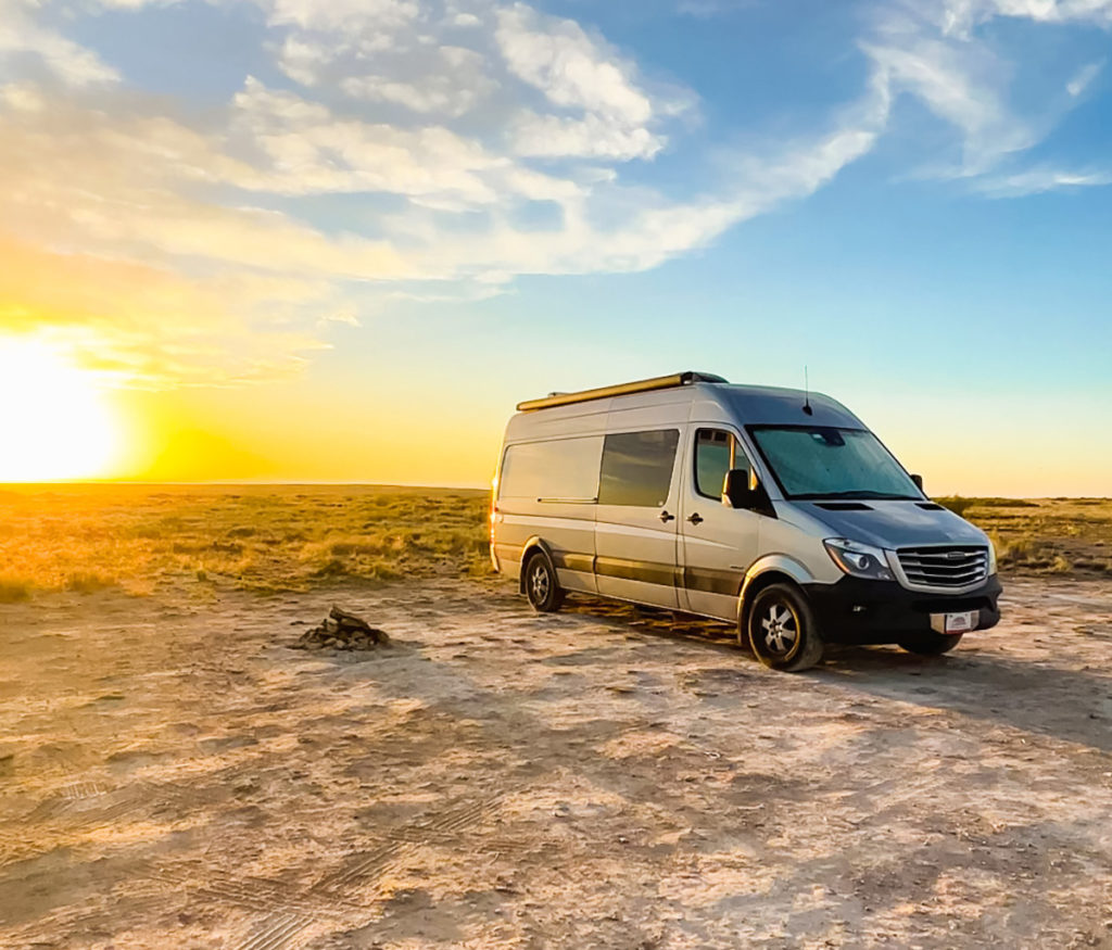 a silver Sprinter campervan is parked next to a fire ring in a desert boondocking (wild camping) spot in Roswell, New Mexico. The sun is setting behind it.