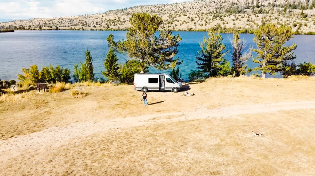 an aerial photo of a woman standing beside her silver Sprinter campervan, which is parked next to a fire ring at a beautiful, massive boondocking (wild camping) spot. Behind the van are a variety of trees, which stand at the shore of a large lake. On the other side of the lake fre hills with scattered trees. this is located in Wyoming.