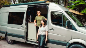 a smiling man and woman stand inside the open sliding side door of their silver Sprinter campervan. some trees are seen in the background.