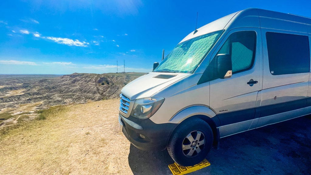 a silver Sprinter campervan sits parked on leveling blocks on the driver side at the edge of an overlook cliff, at a vast boondocking (wild camping) location at the Badlands of South Dakota. it is a sunny day with a few white clouds in the blue sky.