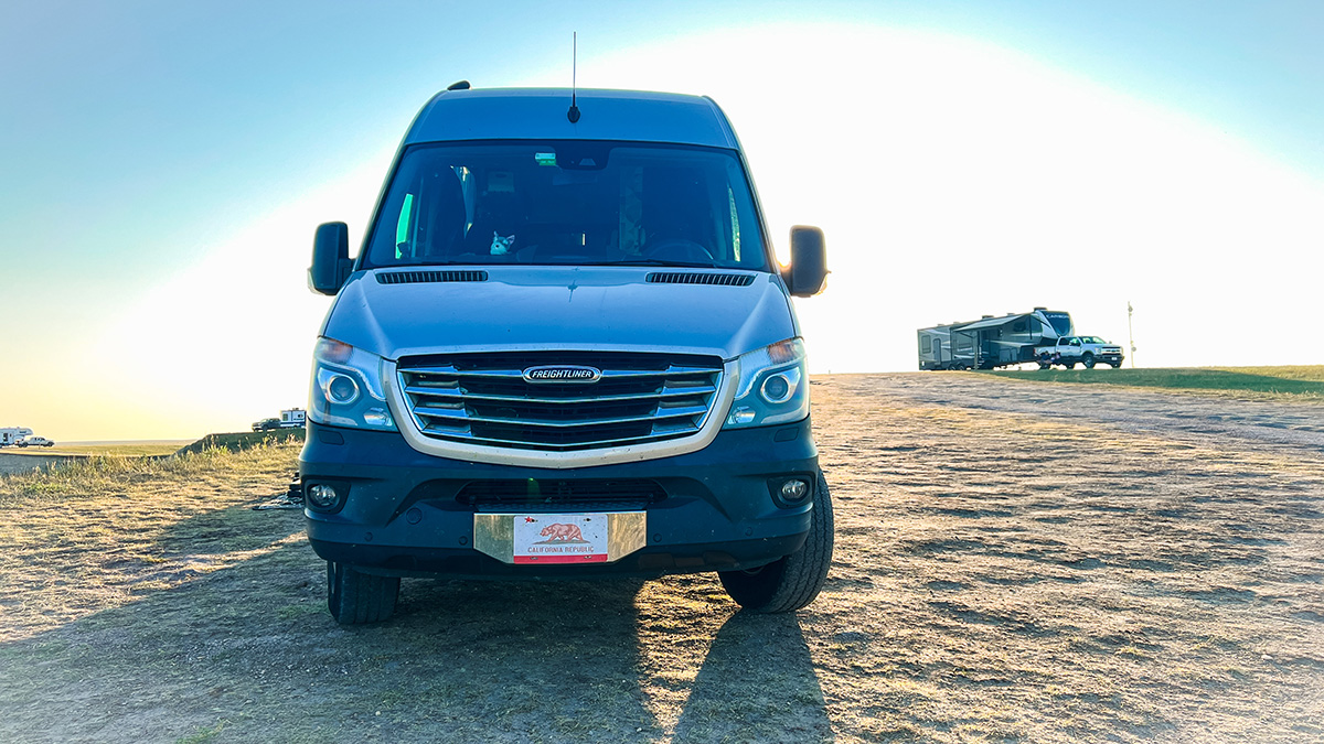 a silver Freightliner Sprinter campervan is parked at a sunny boondocking (wild camping) spot in the Badlands of South Dakota. the van has a California Republic license plate on the front, and in the background are other pickup trucks with fifth wheel campers attached.