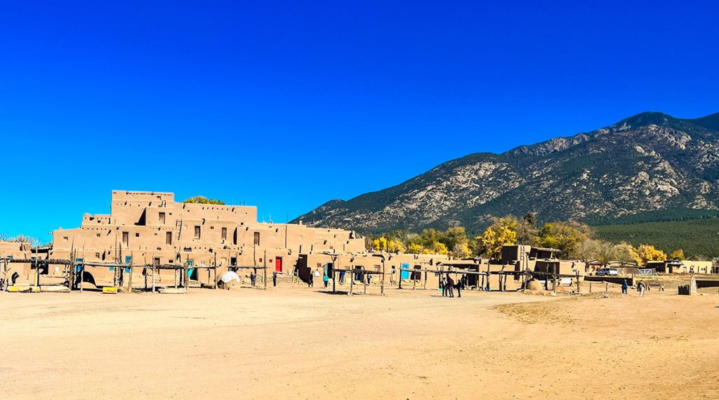 landscape photo of Taos Pueblo multi-storied adobe dwellings with trees and mountainous hill in the background