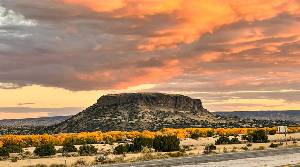 a colorful semi-desert like landscape off the side of a road in New Mexico. A large butte of sorts is in the near distance with the horizon far in the background. dramatic orange and grey clouds fill the sky. the ground is heavily covered with orange-y and dark green bushes.
