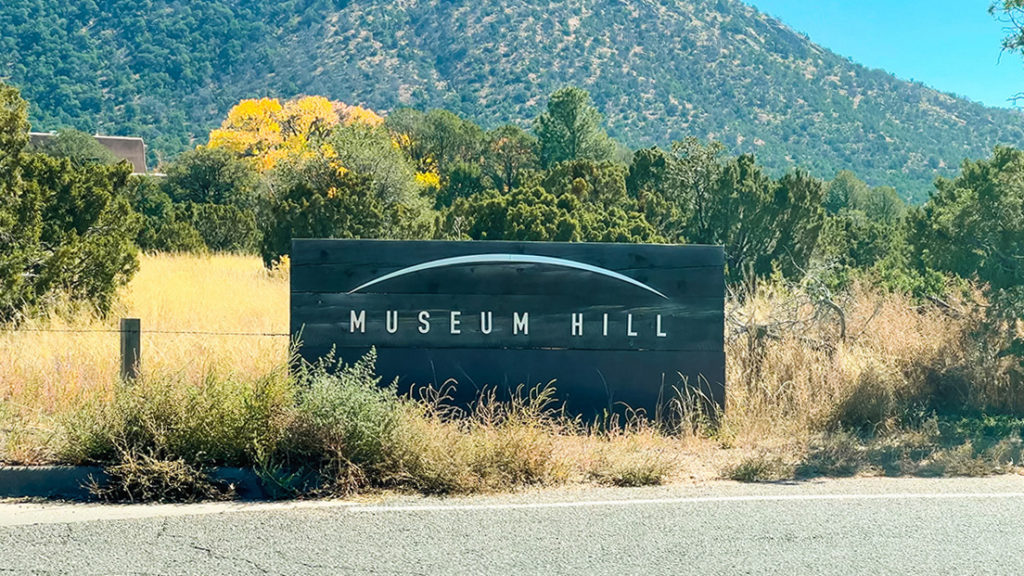 Museum Hill sign with a landscape setting surrounding it, at the entrance to Museum Hill in Santa Fe, New Mexico. a moutainous hill is in the background along with trees and tall grass