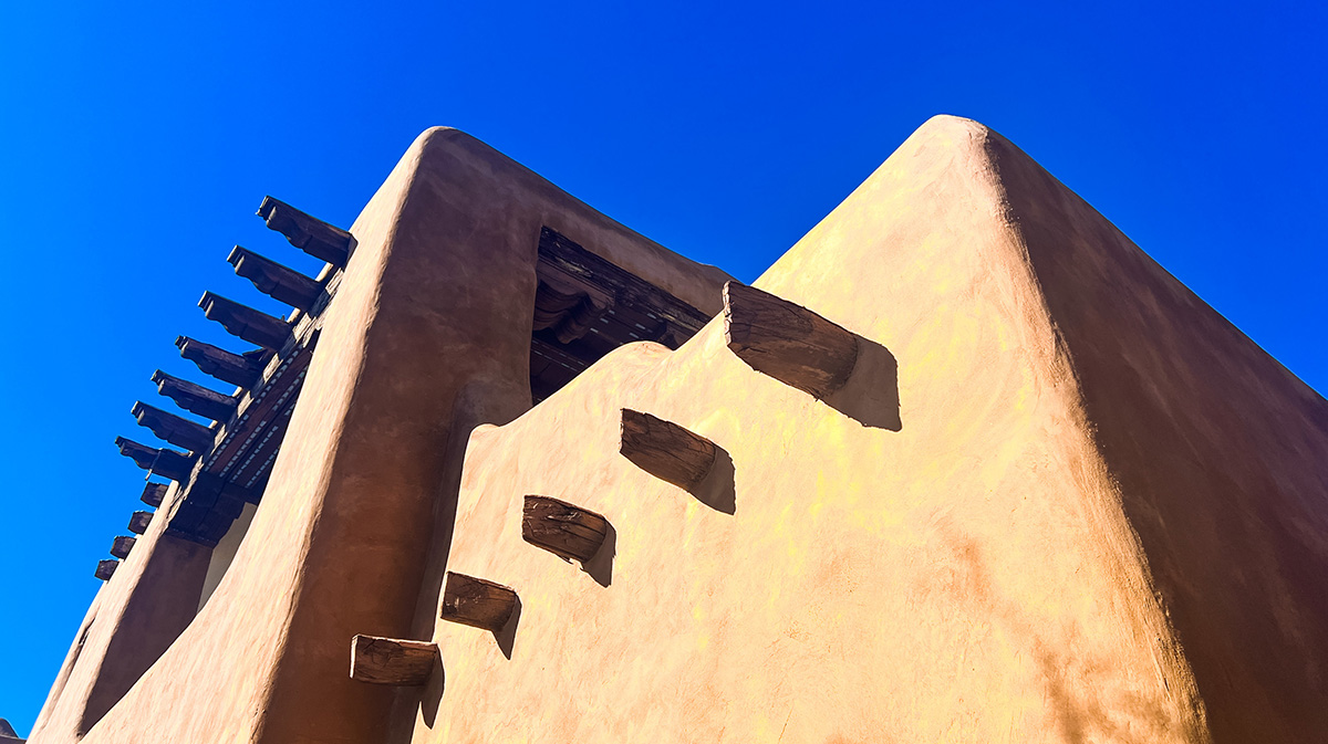 photo of the top of an adobe building in Santa Fe, New Mexico, shot from the ground up. visible are corners of the roof and several of the traditional wooden beams used in this construction