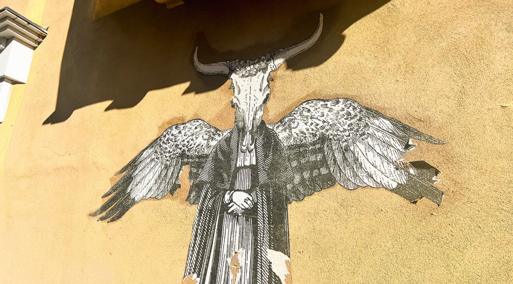 a painting on the side of an adobe building in Santa Fe, New Mexico depicts a person in a robe and cloak, with large wings and a longhorn steer skull for a head
