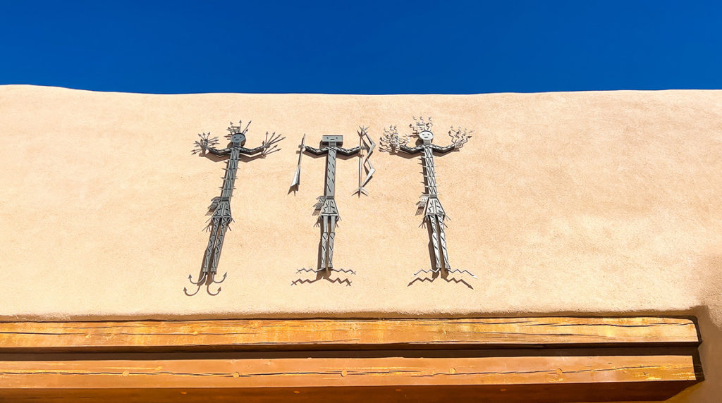 art sculptures at the top of an adobe building in Santa Fe New Mexico, which is part of one of the museums on Museum Hill. Native in style, it features three people, each holding different items such as bow and arrows