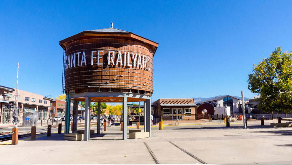 a scene at the Railyard in Santa Fe, New Mexico. a large elevated water tank with "Santa Fe Railyard" and a ladder on it sits next to railroad tracks and in the foreground, with several buildings in the background. a large flying saucer sculpture can be scene on its side, leaning against one of the buildings