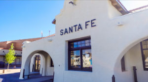 a white adobe style building with letters reading "Santa Fe" over a window, and arched entry ways on either side. Santa Fe, New Mexico.