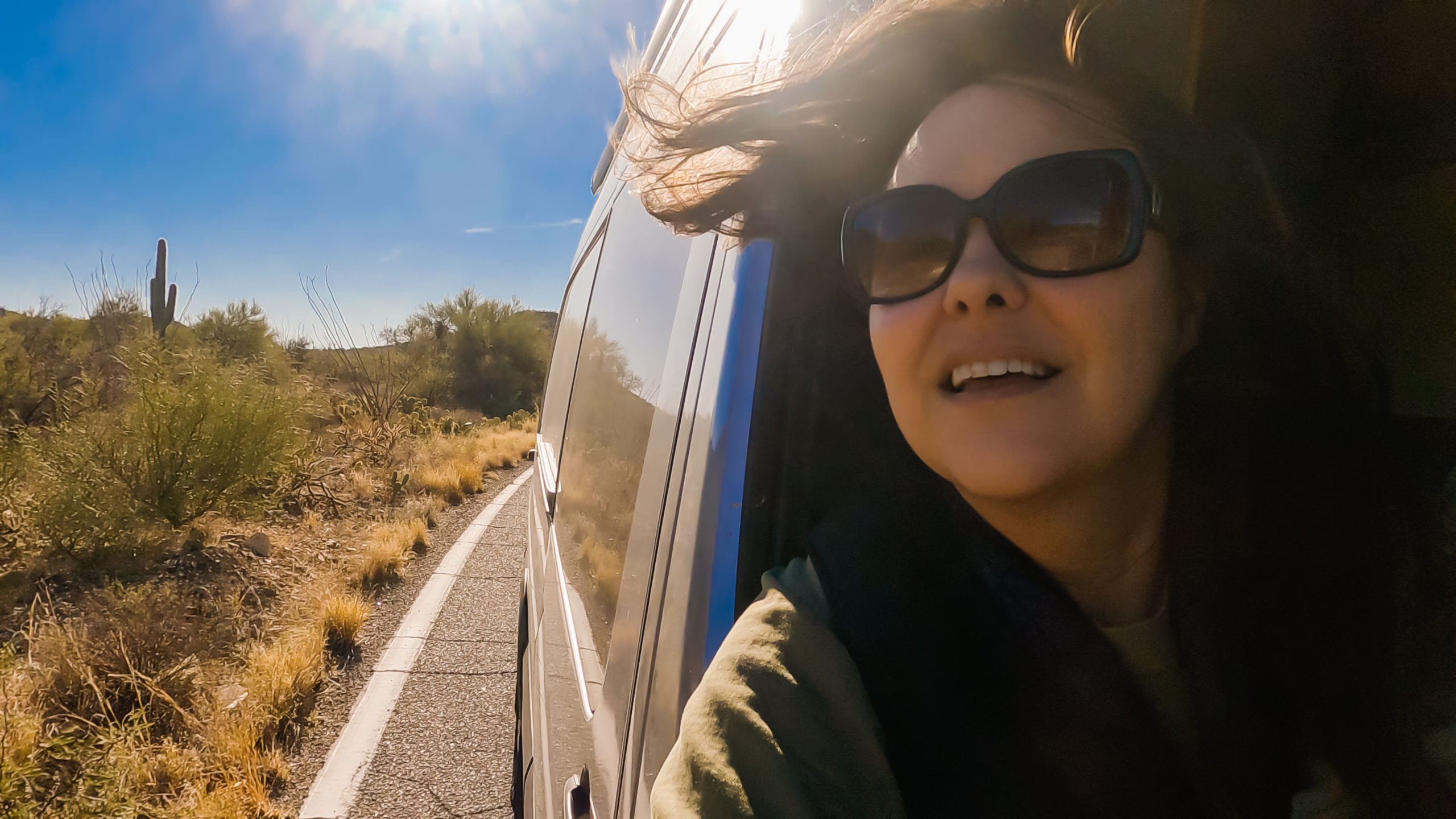 woman with her head out the passenger window ofa campervan, wind blowing her hair, with the road and cacti in the background. road trip apps